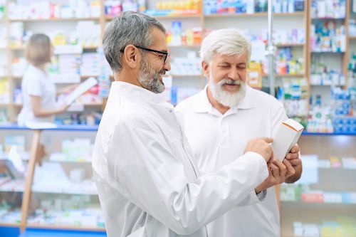 Two pharmacists examining medication in a pharmacy. One wearing glasses smiles, other has a beard.