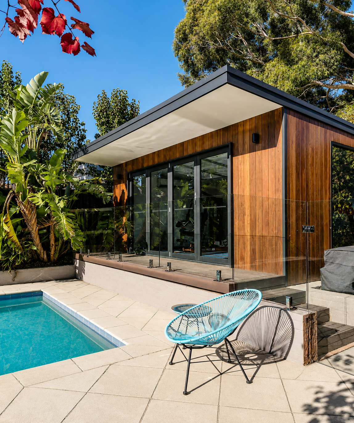 Modern poolside patio with glass doors, wood siding, and a turquoise lounge chair under a blue sky