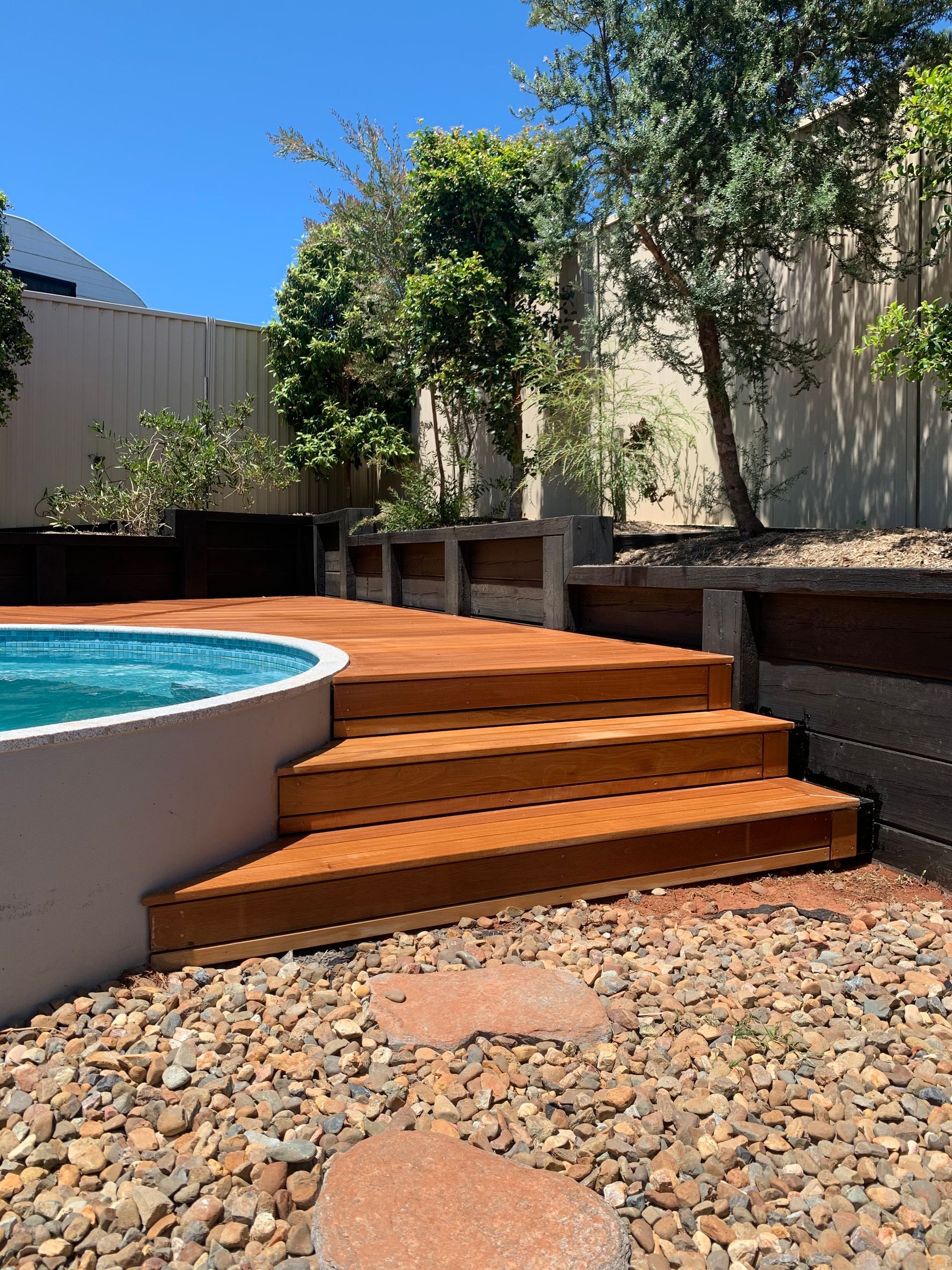 Backyard pool with wooden steps, raised deck, and gravel landscaping under bright sunlight