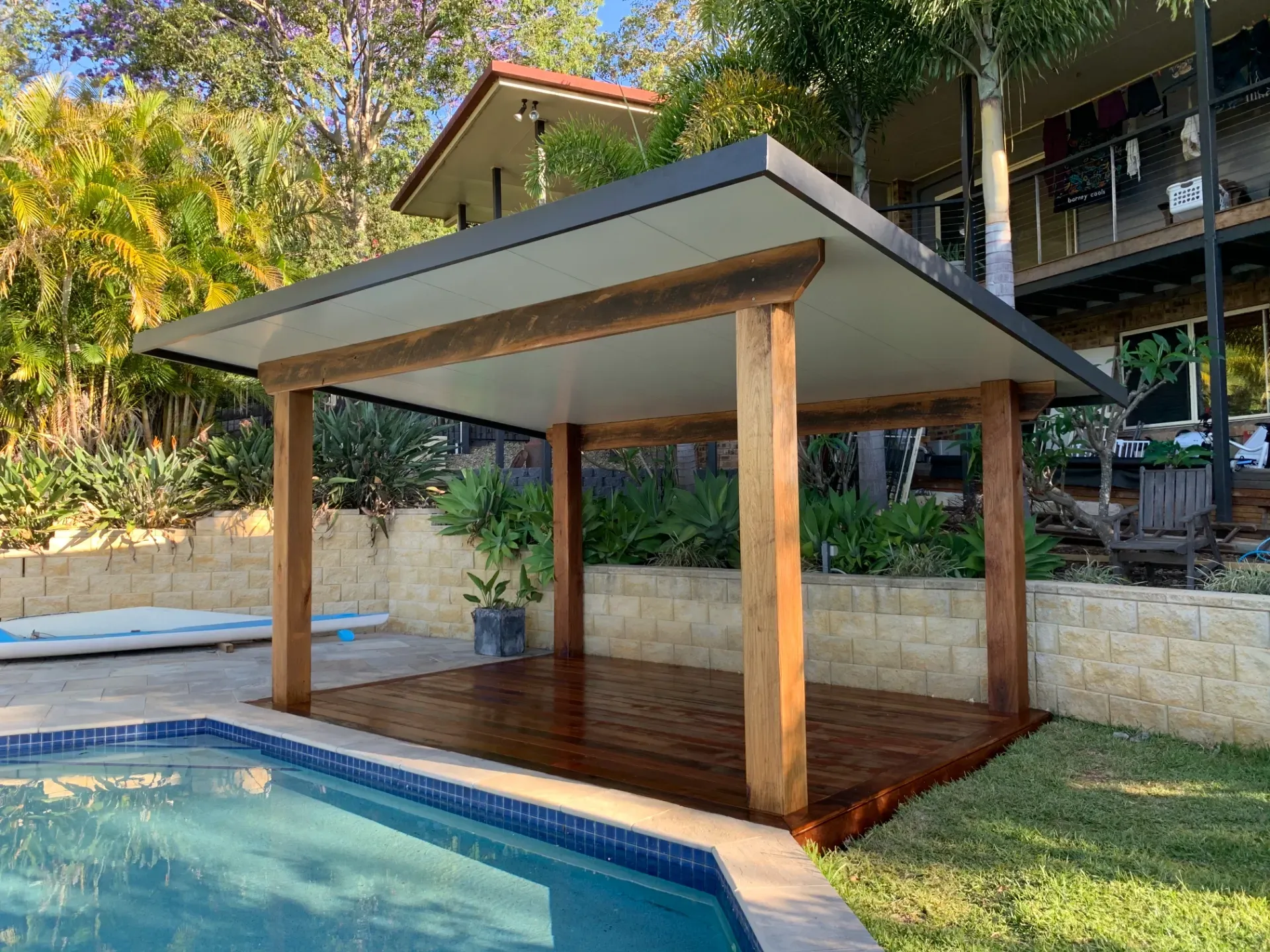 Covered wooden poolside pavilion with a gray roof beside a swimming pool and tropical landscaping