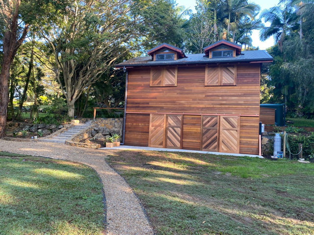 Wooden house with two dormer windows, surrounded by trees and a grassy yard with a curving path.