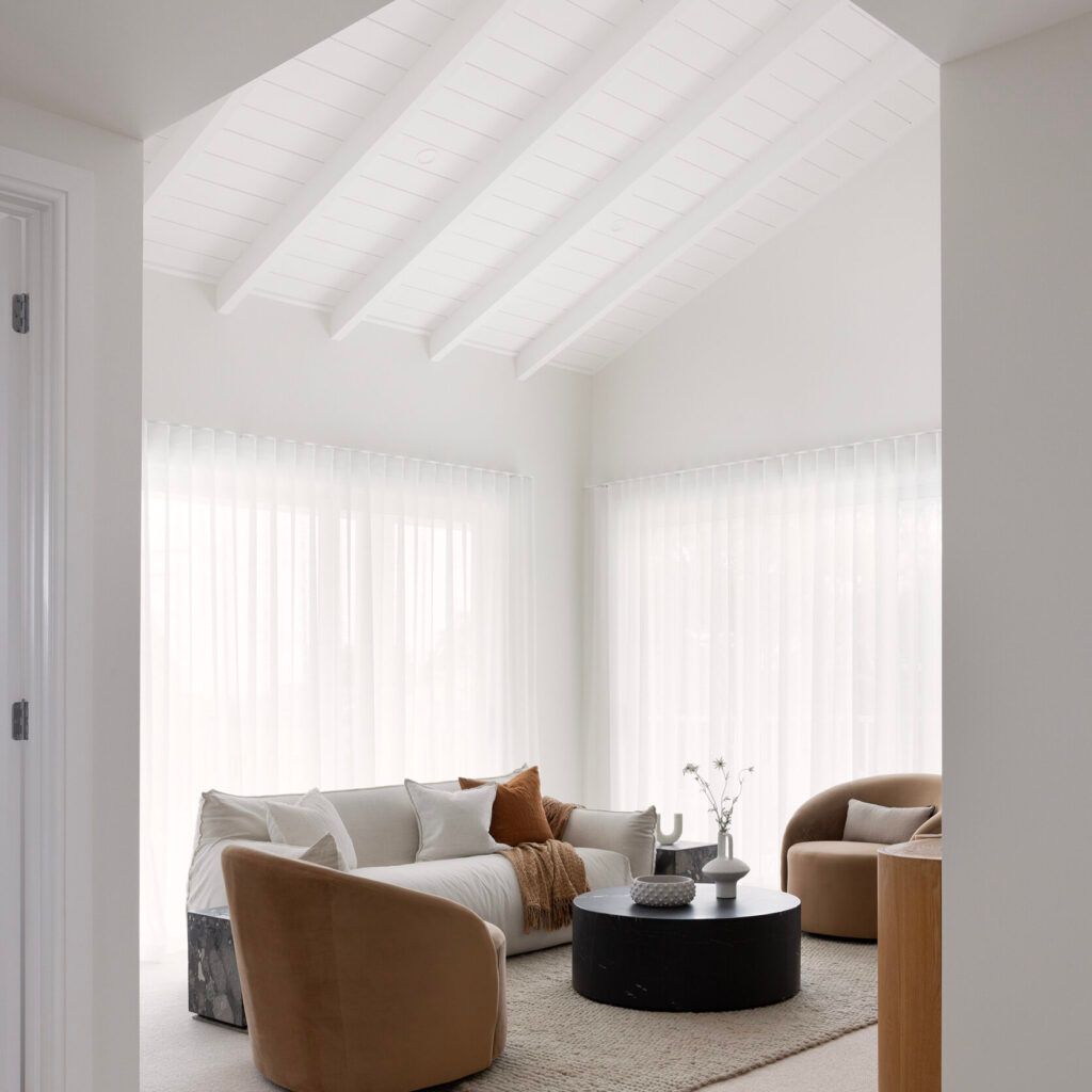 Bright white modern living room with vaulted ceiling, sheer curtains, beige chairs, and round black coffee table
