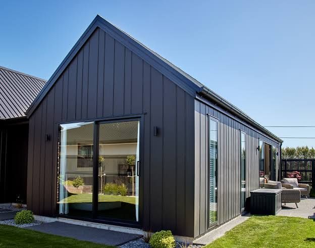 Modern black house exterior with large windows and a small patio in a sunny yard