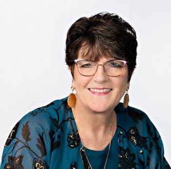 Picture of Cyndi in a blue jacket with black and gold designes on it. Cyndi weares glasses and has on gold earrings.