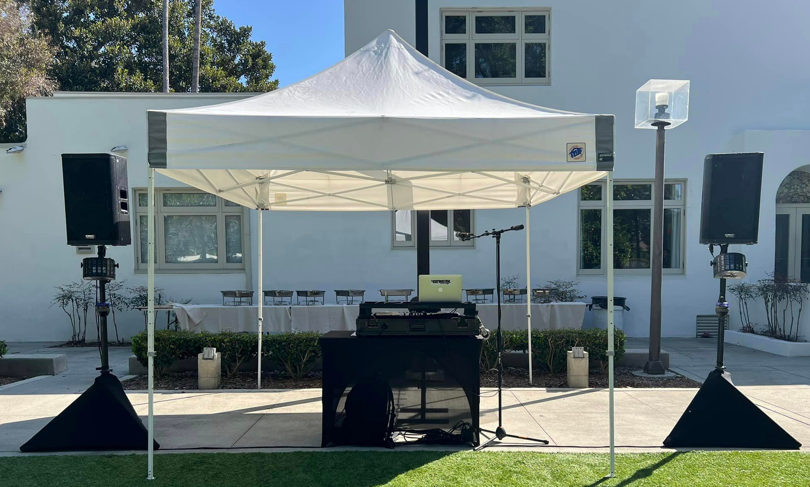 DJ setup under white canopy; speakers, equipment, and microphone stand in front of a building.