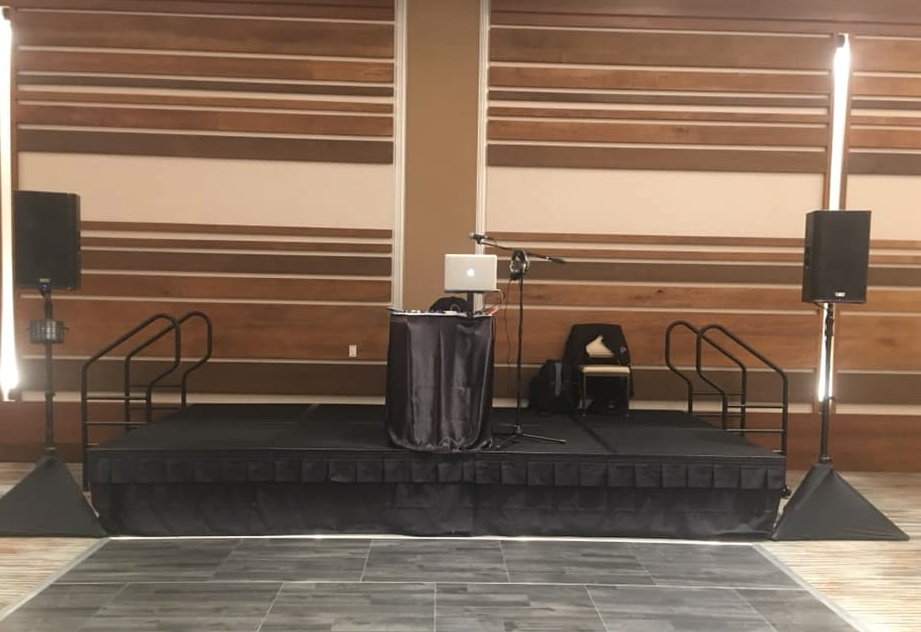 A stage with black facade, DJ setup, speakers, and a laptop, set in a venue with wood paneling.