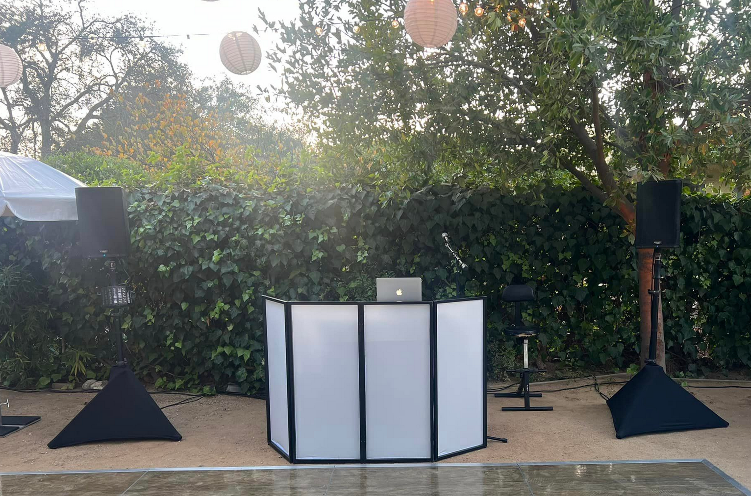 DJ setup outdoors: white DJ booth, speakers, microphone, laptop on a patio with trees and hanging lanterns.