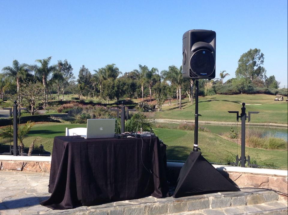 DJ setup outdoors on a patio overlooking a golf course. Black table, laptop, speaker on stand, blue sky.