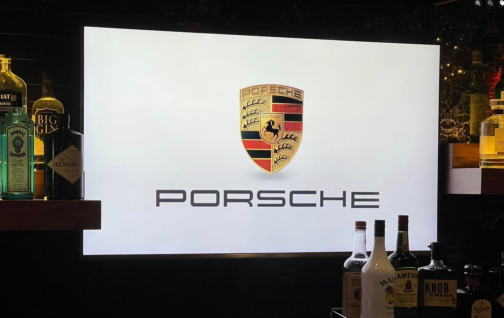 Porsche logo on a white background in a bar, with bottles of liquor visible.