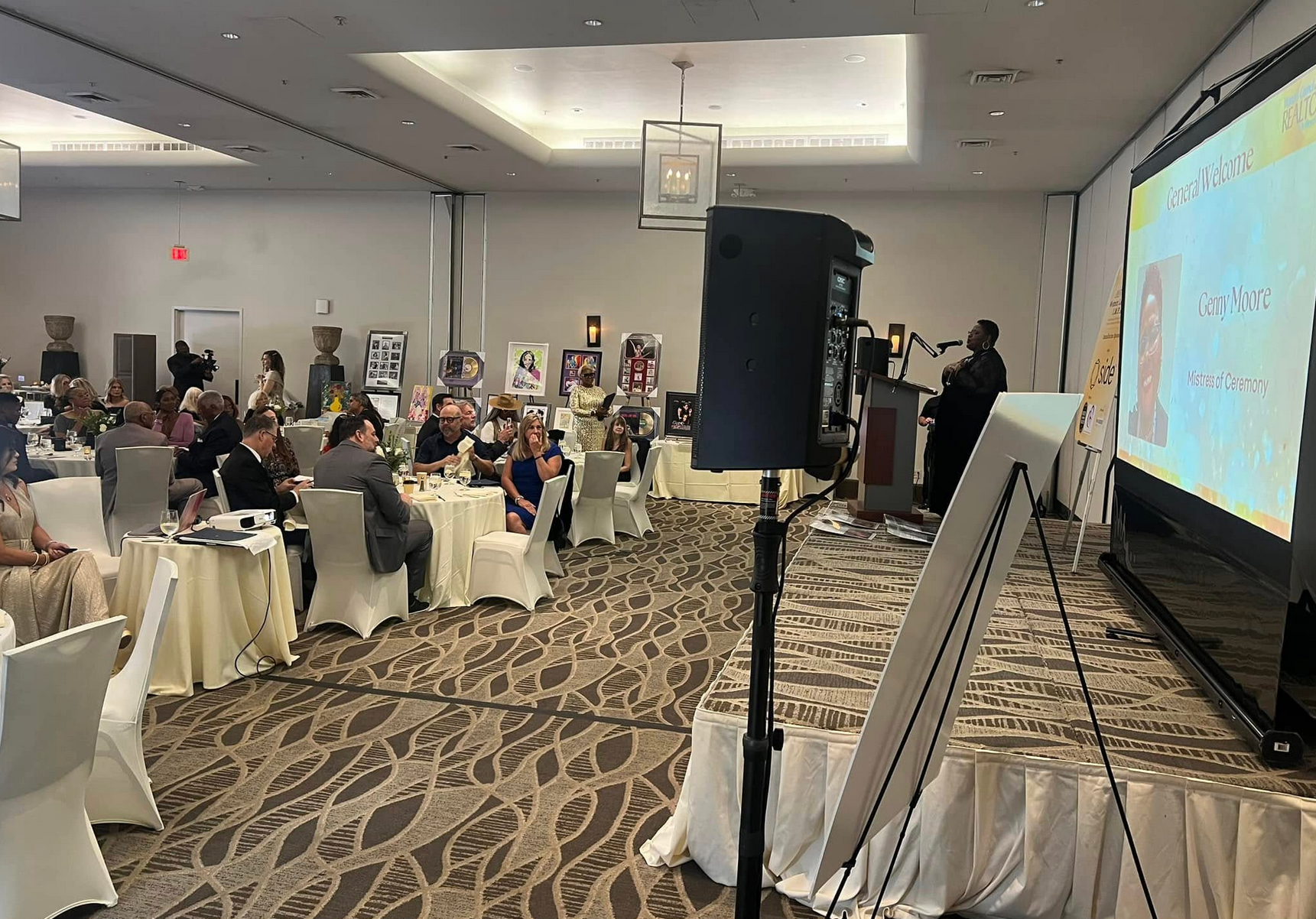 Man giving a speech at an event with people seated at tables. A projector displays images.