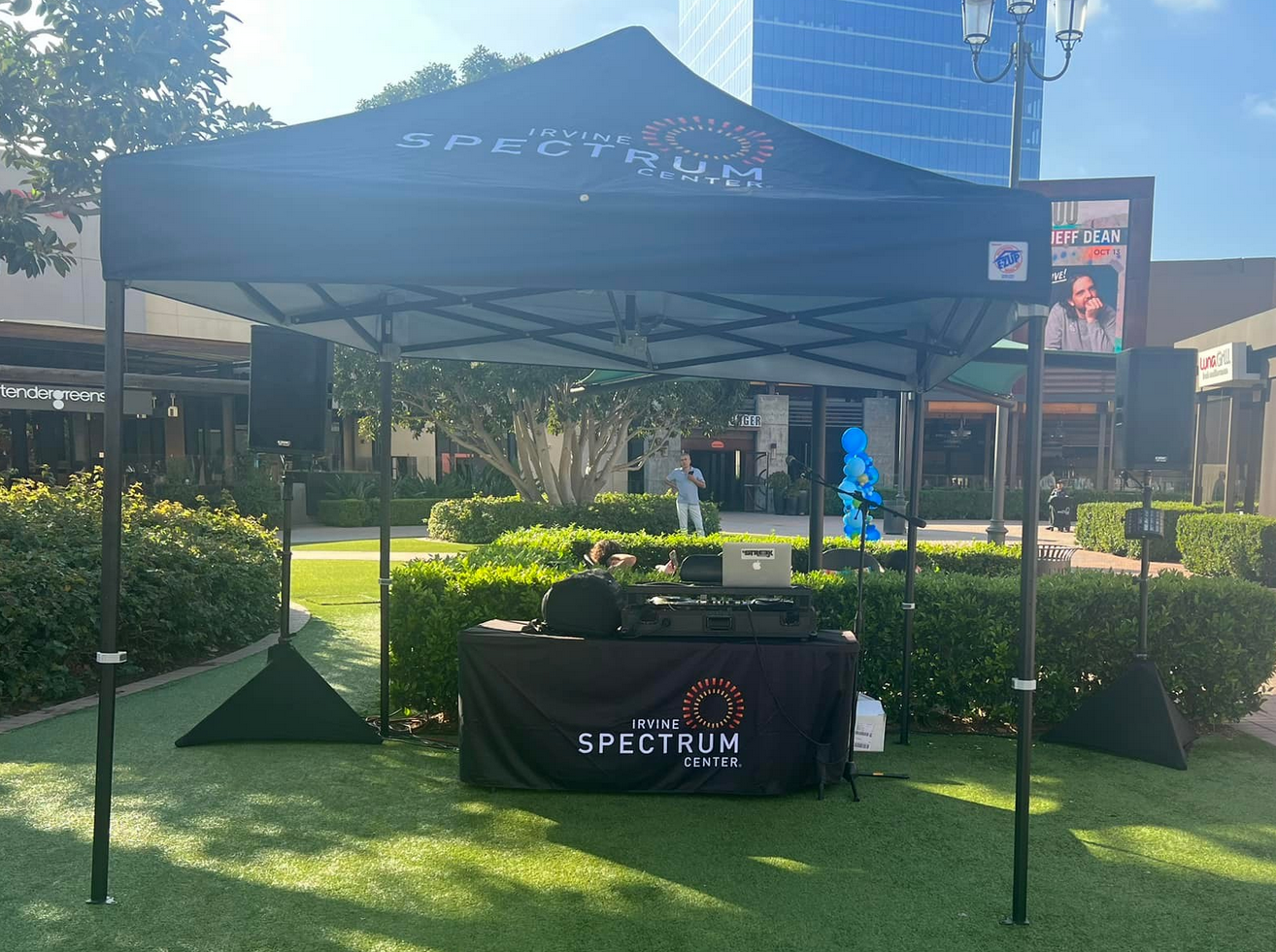 DJ booth setup under a black canopy on green grass; building and trees in background.