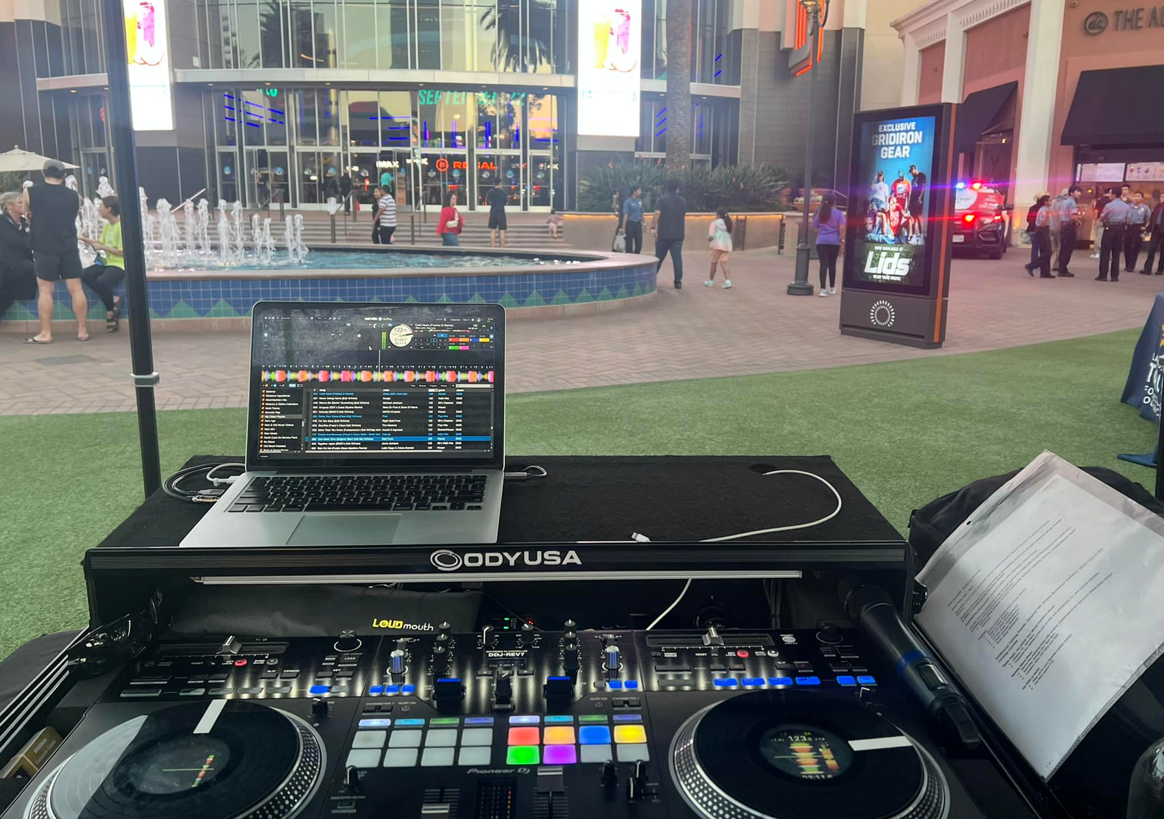 DJ setup outside: laptop, mixer, and turntables with a fountain and people in the background.