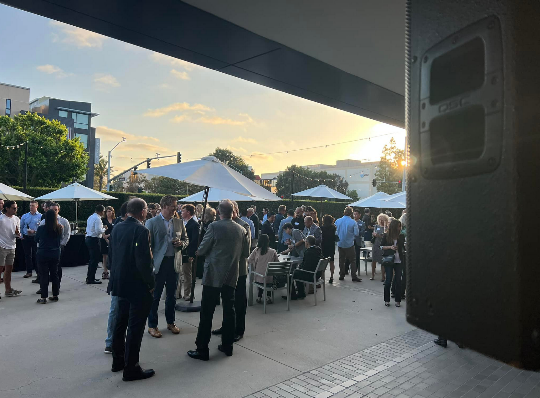 Group of people at an outdoor event on a patio during sunset. Some standing, some at tables.