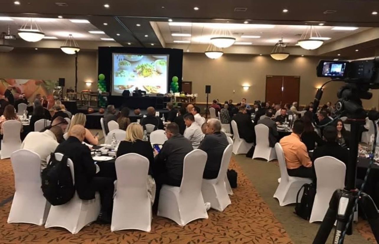 Large banquet hall with many people seated at tables; screen at front; video camera in foreground.