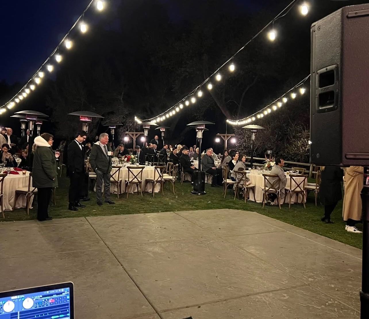 Outdoor evening event; tables set with guests, string lights, heaters, speaker, and DJ setup.