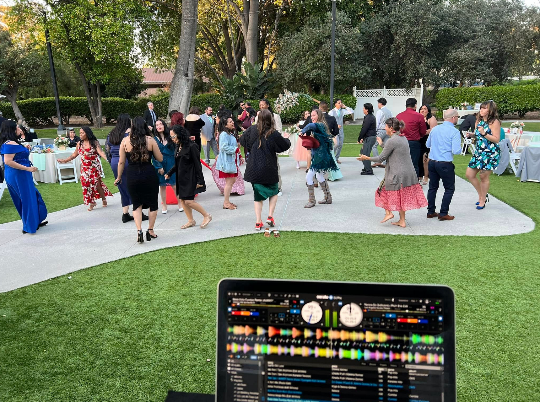 People dancing at an outdoor event with a DJ setup in the foreground, green lawn and trees.