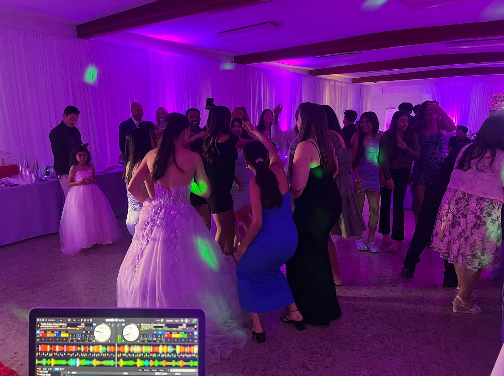 People dancing at a party with purple lighting. DJ equipment in the foreground.