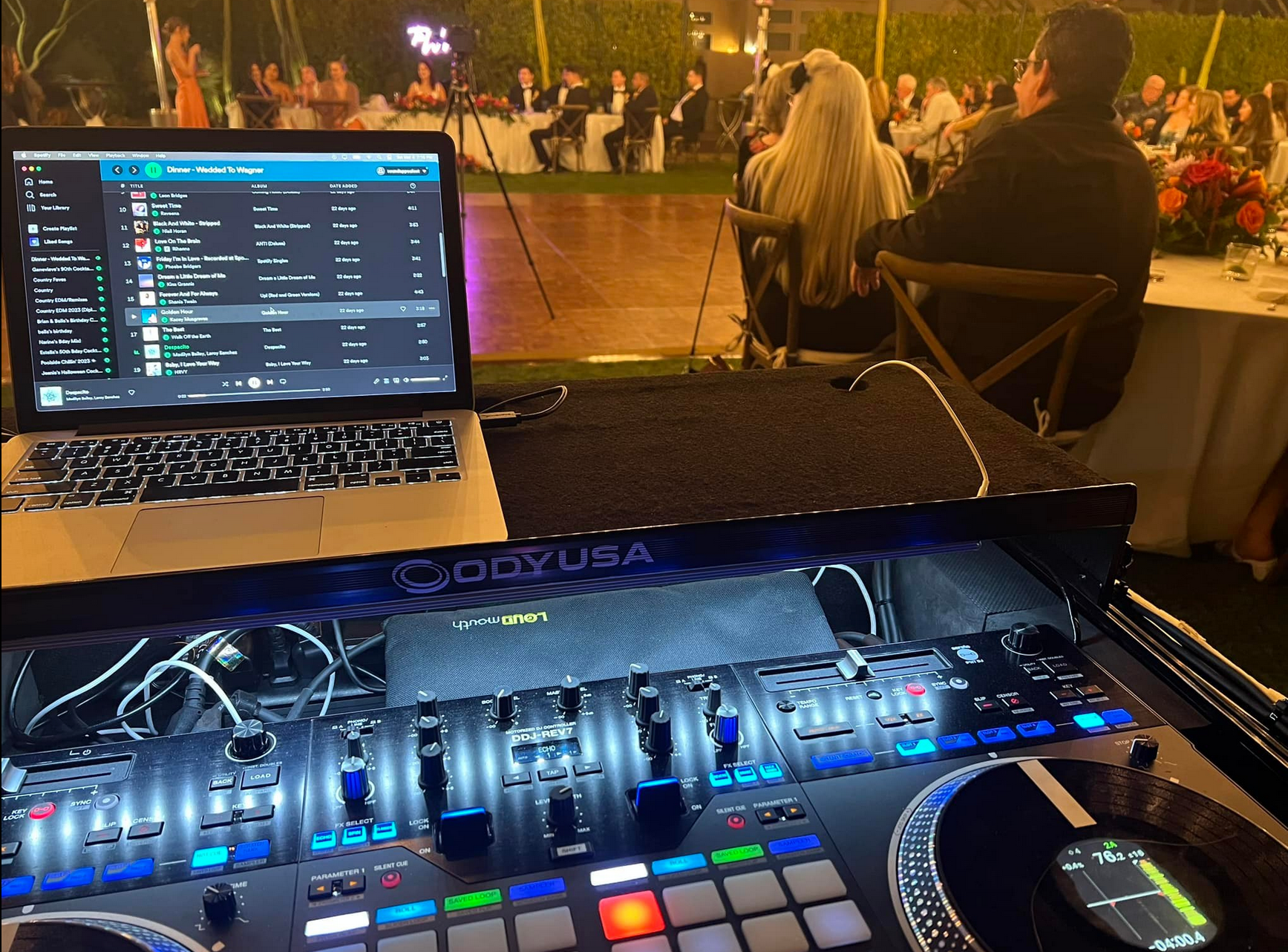 DJ setup with laptop, mixing board, and speakers at an outdoor event with guests seated at tables.