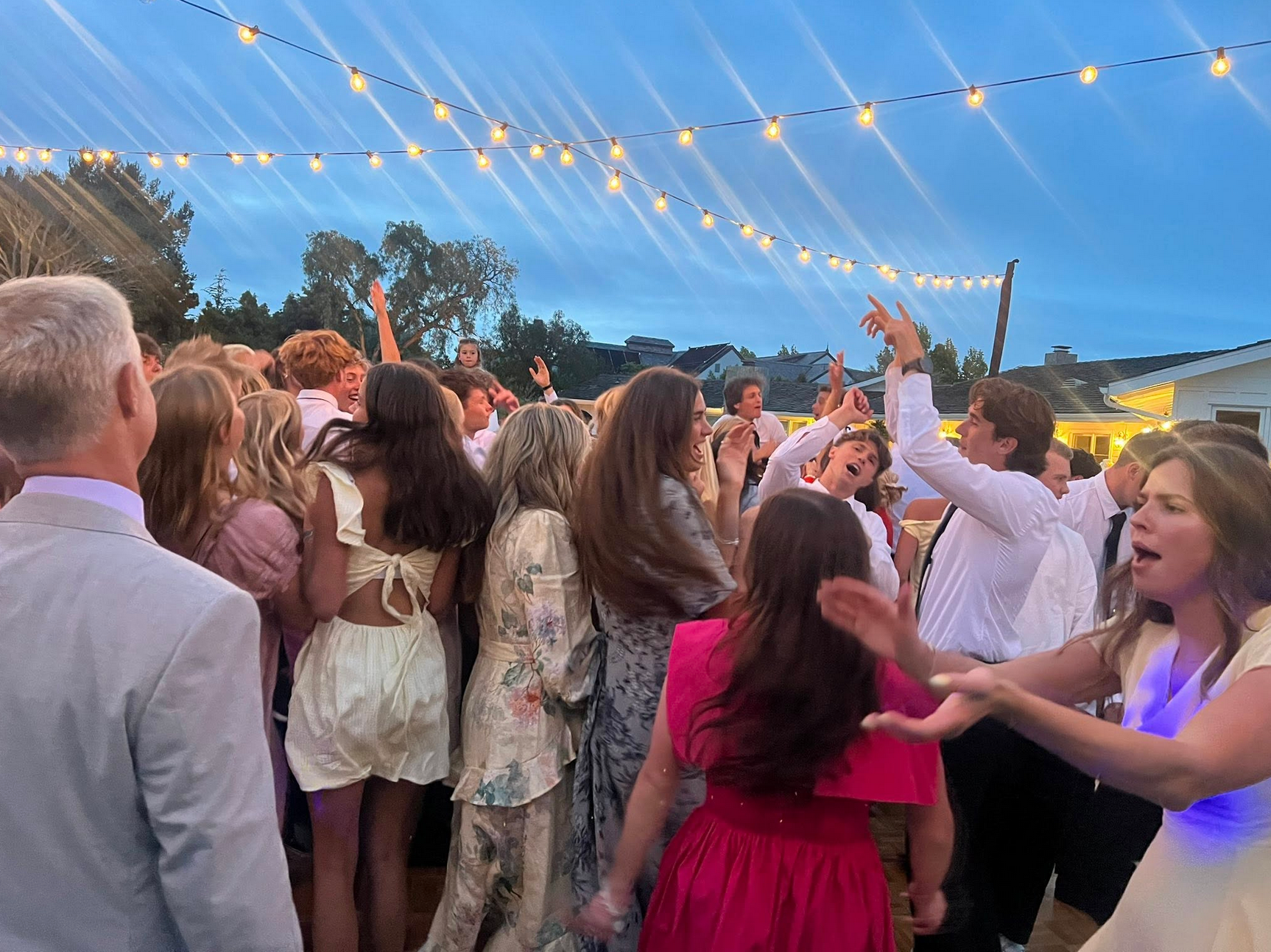 A crowd dances under string lights outdoors; people celebrate, arms raised; evening setting.