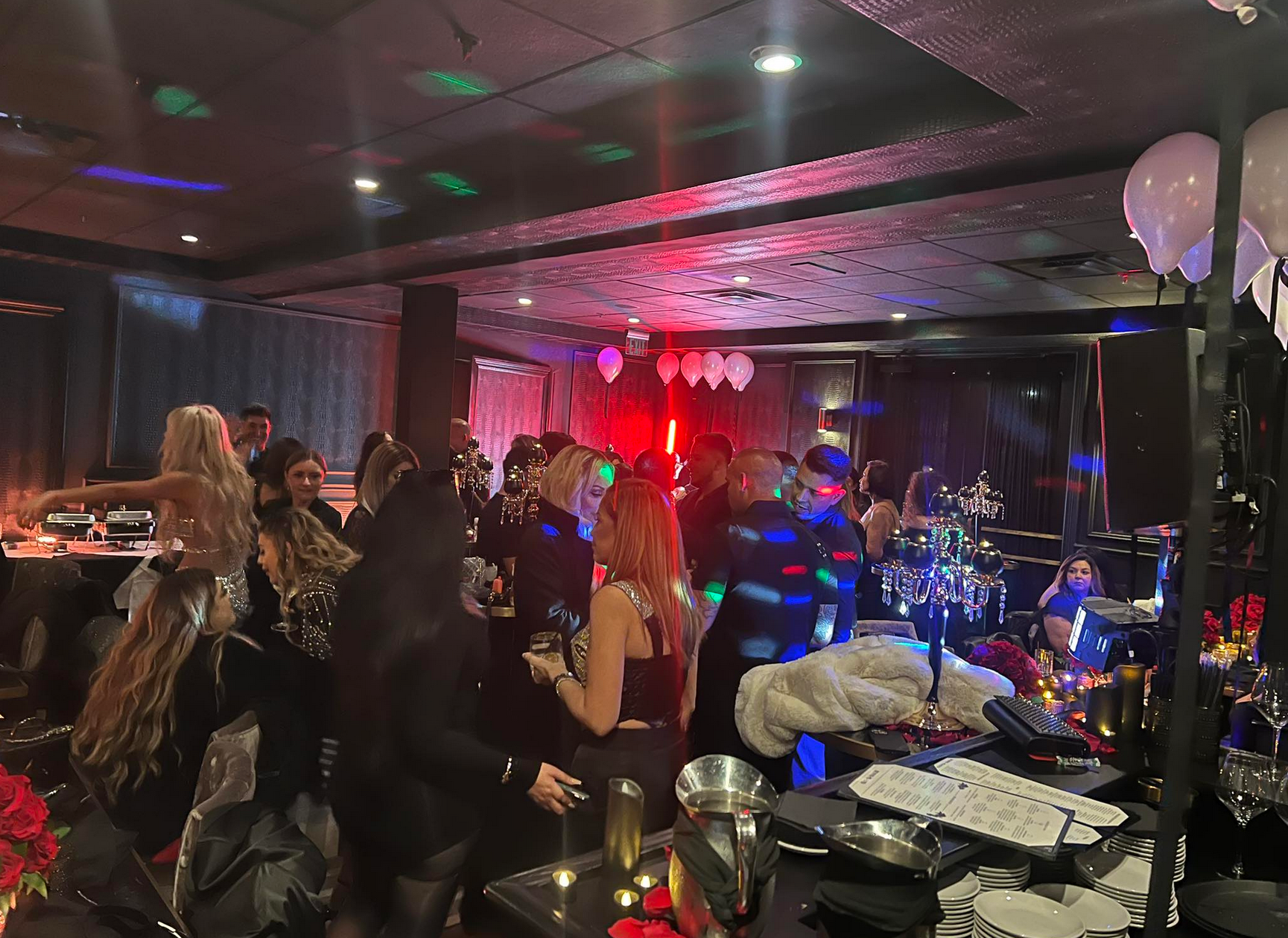 People dancing at a party in a dimly lit venue with disco ball lights, balloons, and red accents.