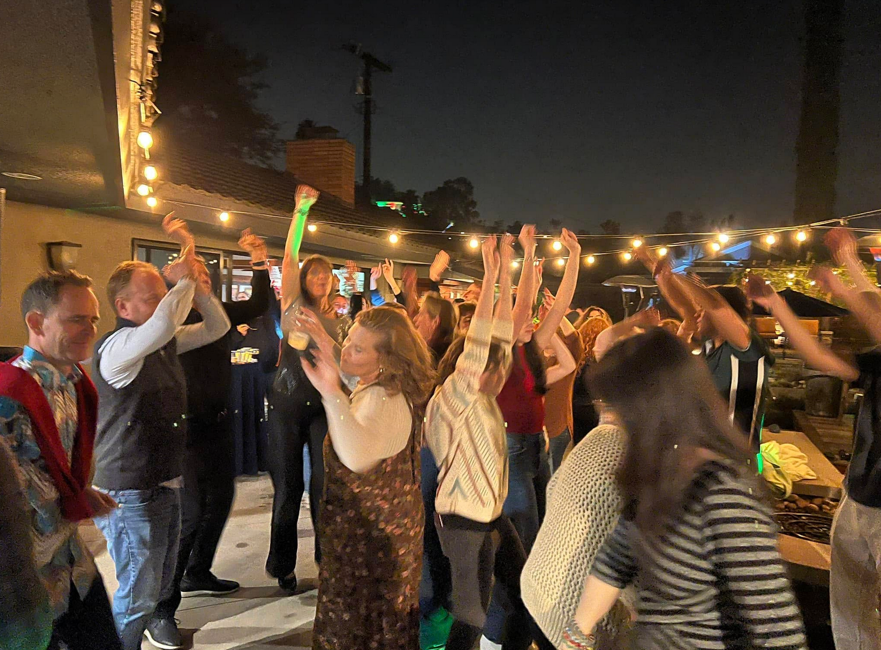 People dancing and raising hands outdoors at night, illuminated by string lights.