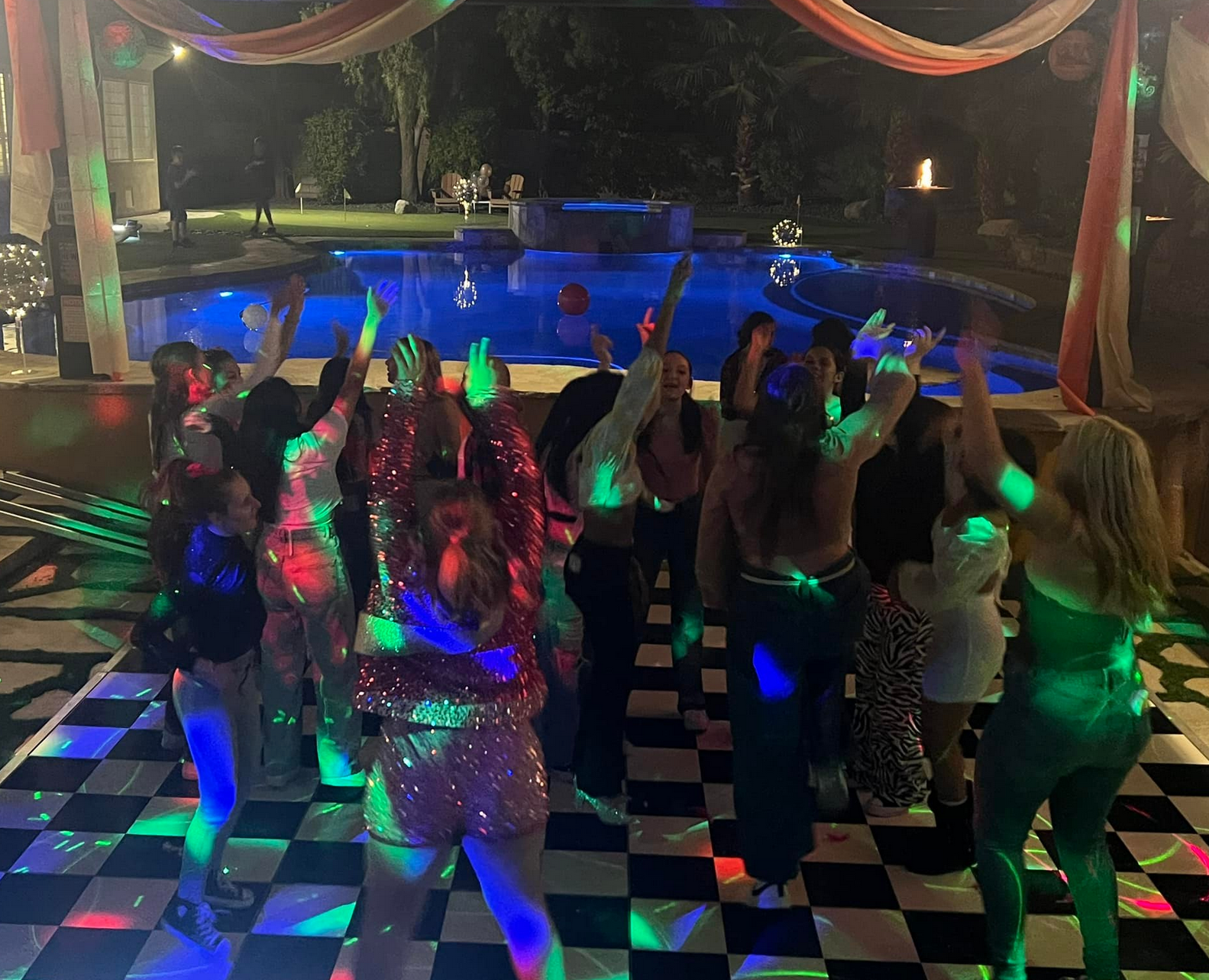 People dancing on a checkered floor near a pool at night, with colorful lights.