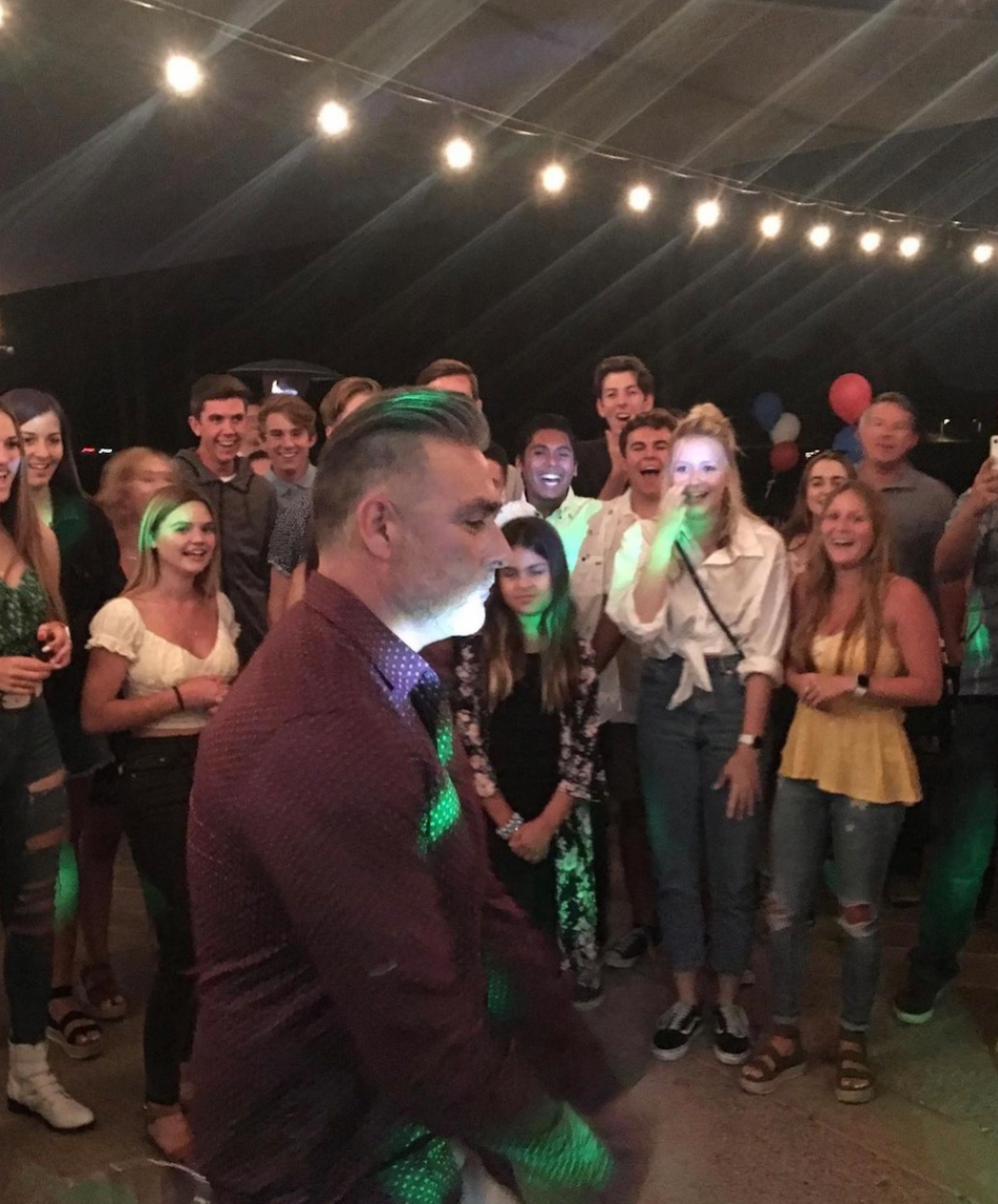 Group of people gathered indoors, watching a man sing into a microphone, lit by string lights.