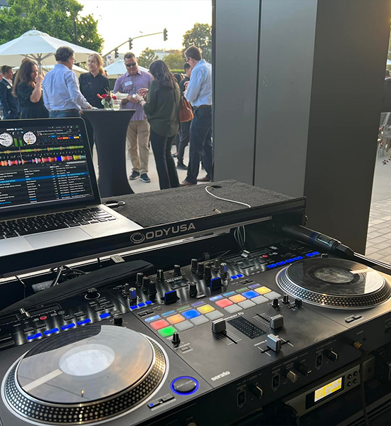 DJ setup with laptop, turntables, and mixer; people socializing in the background outdoors.
