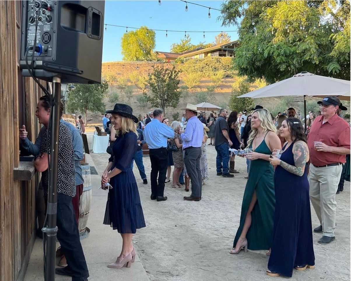 Guests at outdoor event; people in casual attire near a wooden bar and under umbrellas.