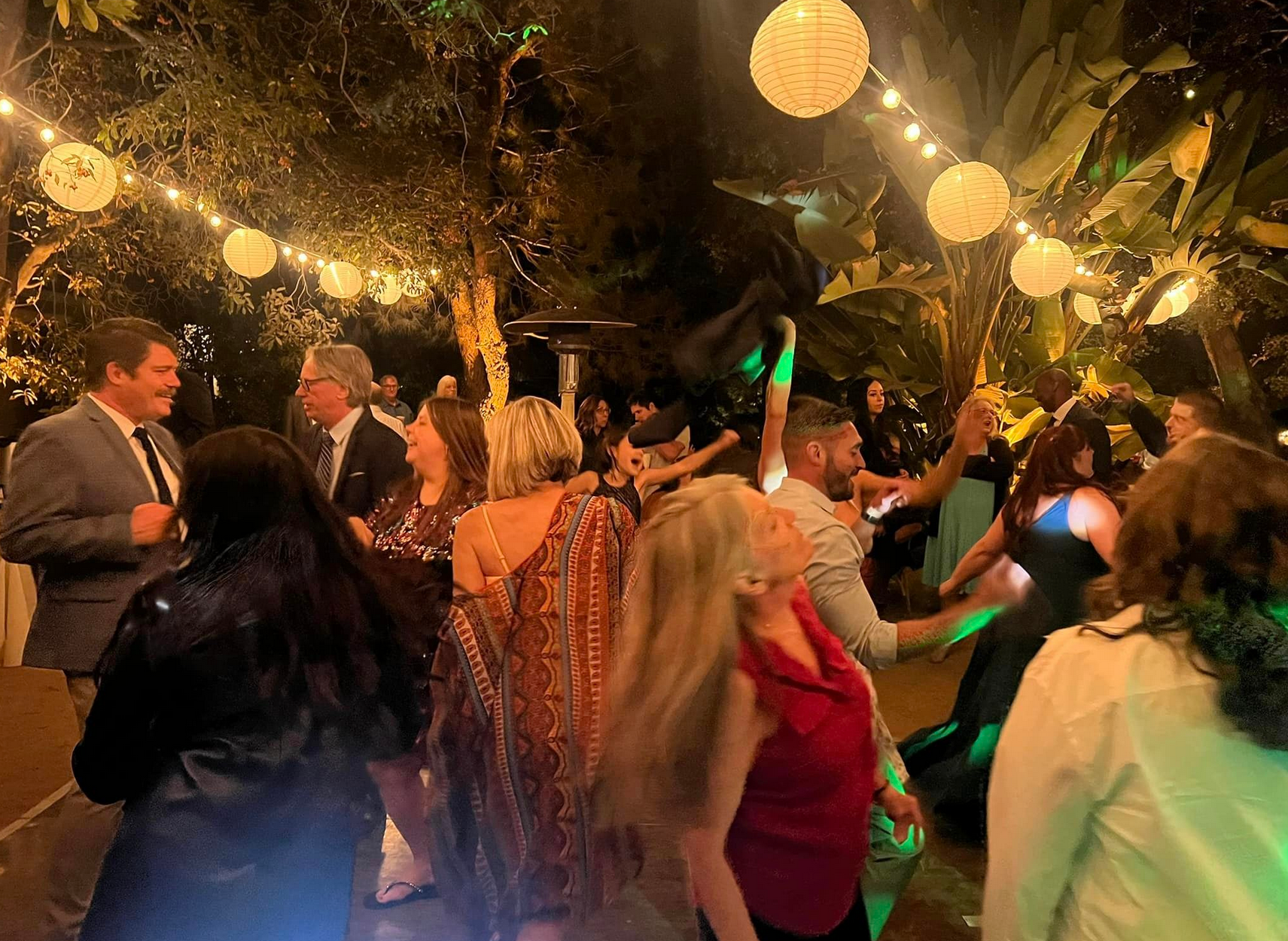 People dancing at an outdoor party with string lights and greenery at night.