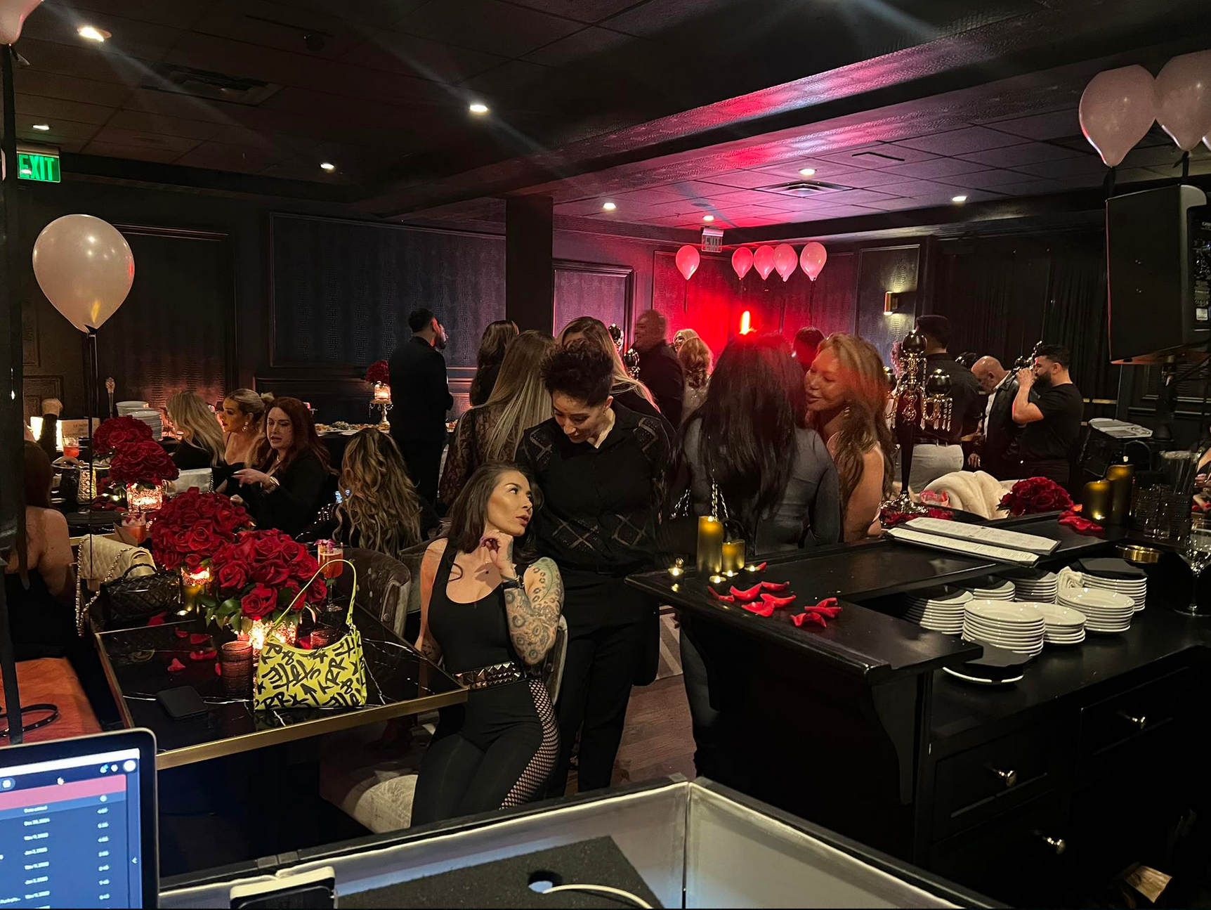 People at a dimly lit party with red flowers, balloons, and a bar.
