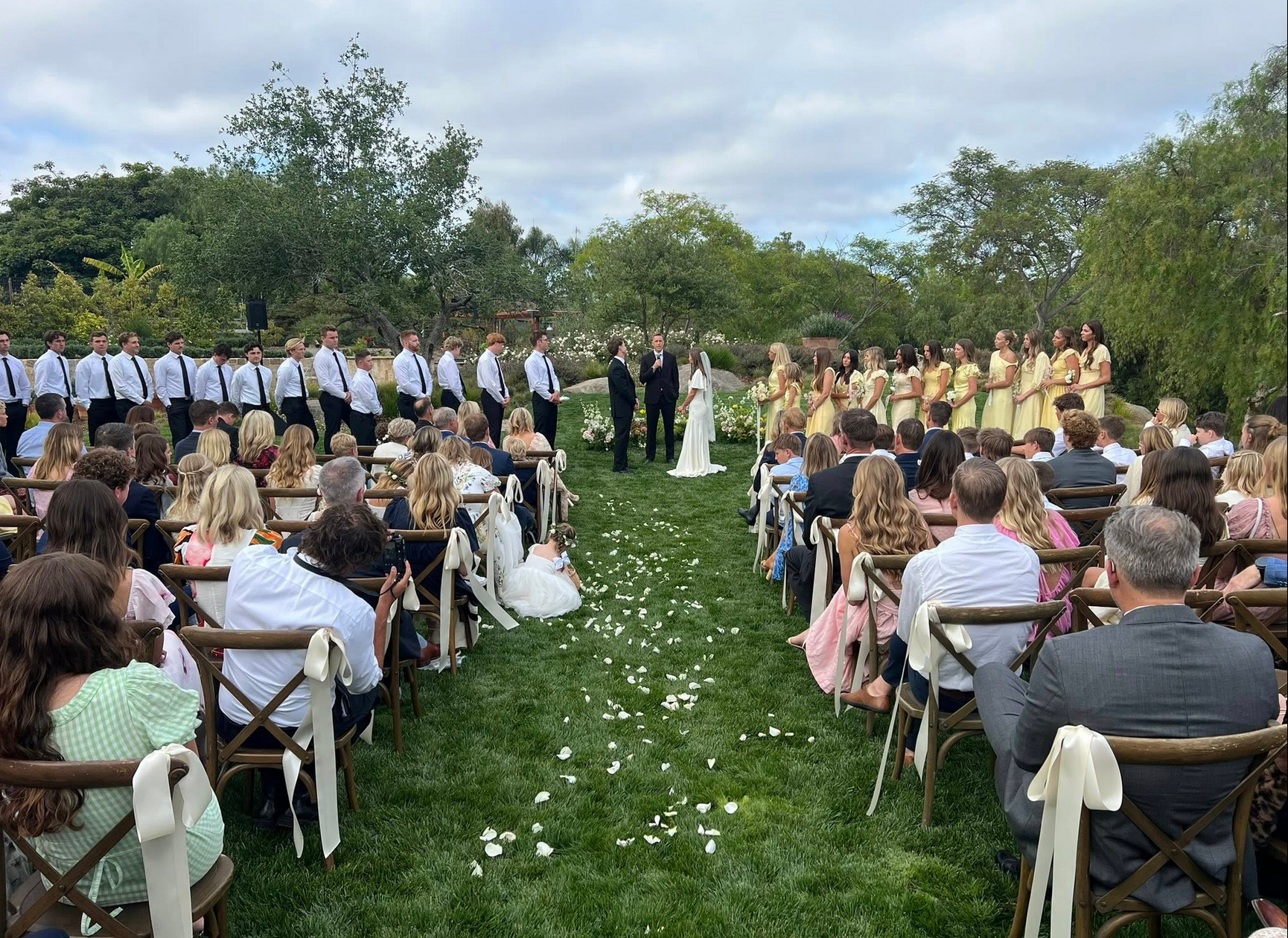 Wedding ceremony in a garden: Bride and groom at altar, wedding party, guests seated on both sides.