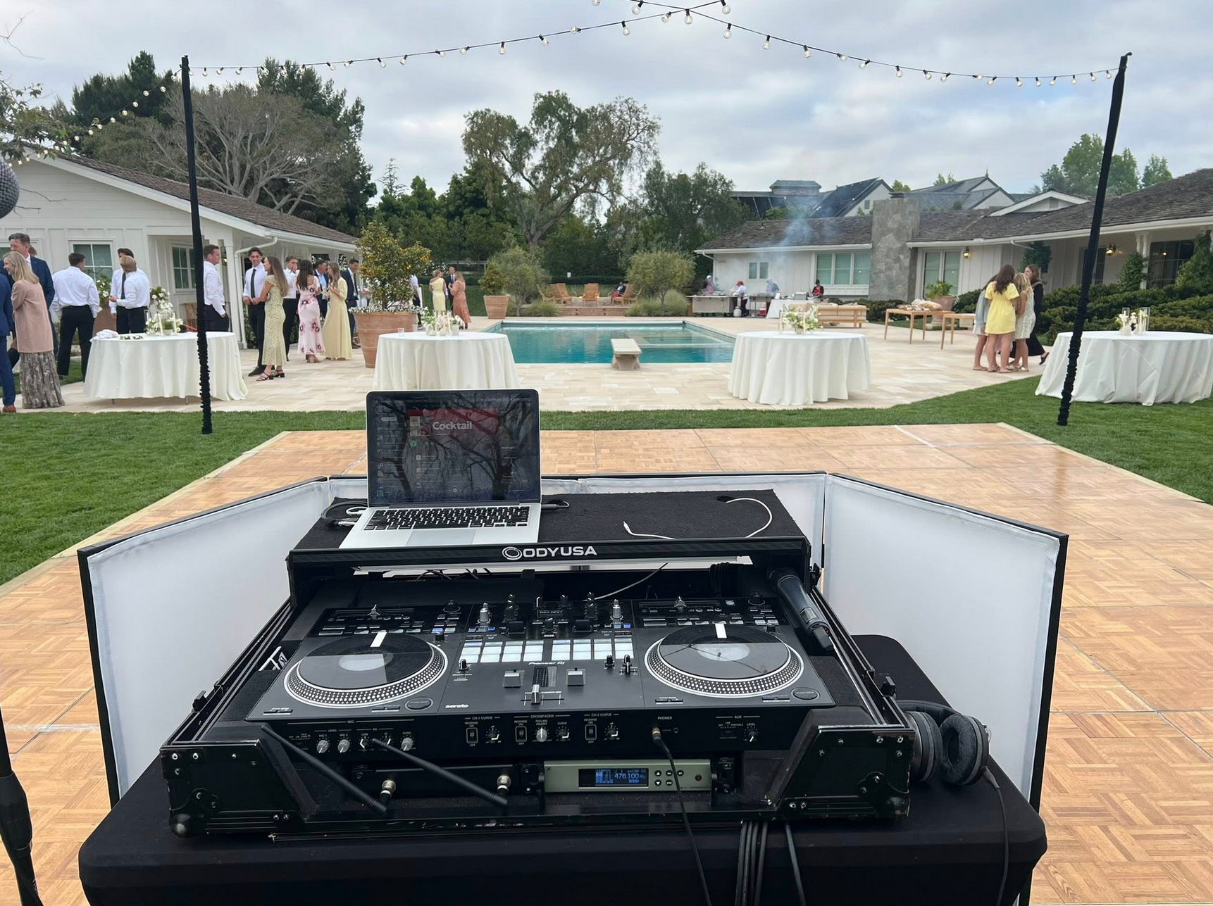 DJ setup at outdoor event, with a pool, white tables, and guests in the background.
