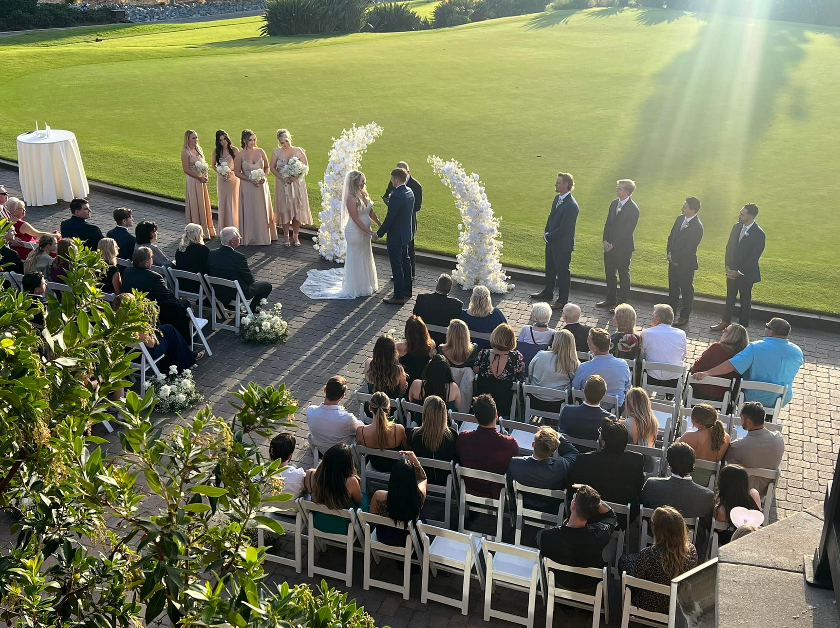 Wedding ceremony outdoors: Bride, groom, bridal party, and guests on a sunny lawn.