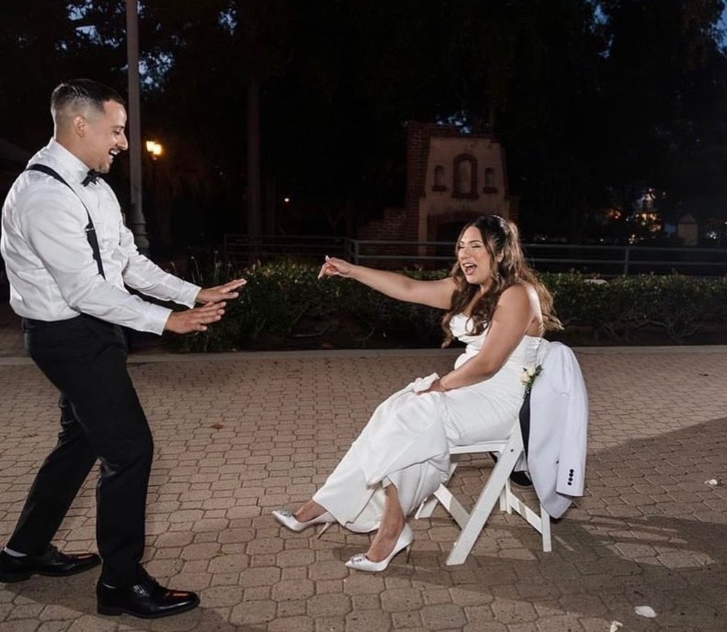 Groom dances towards bride seated on a chair, pointing, in a park at night. She wears white dress and laughs.