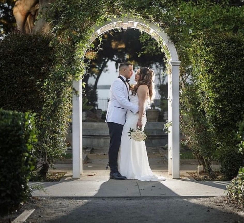 Bride and groom embrace under an archway draped with greenery, outdoors, sunlight.