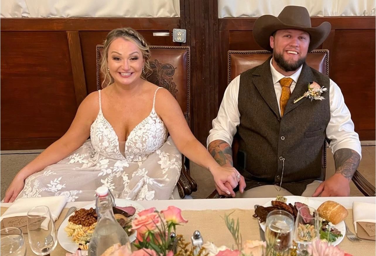 Bride and groom at wedding reception, seated, holding hands, smiling. She in white dress, he in vest and cowboy hat.