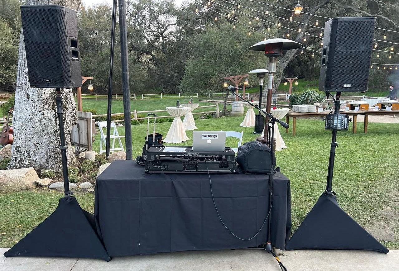 DJ setup outdoors with speakers, mixer, laptop, and microphone on a table covered with a black cloth.