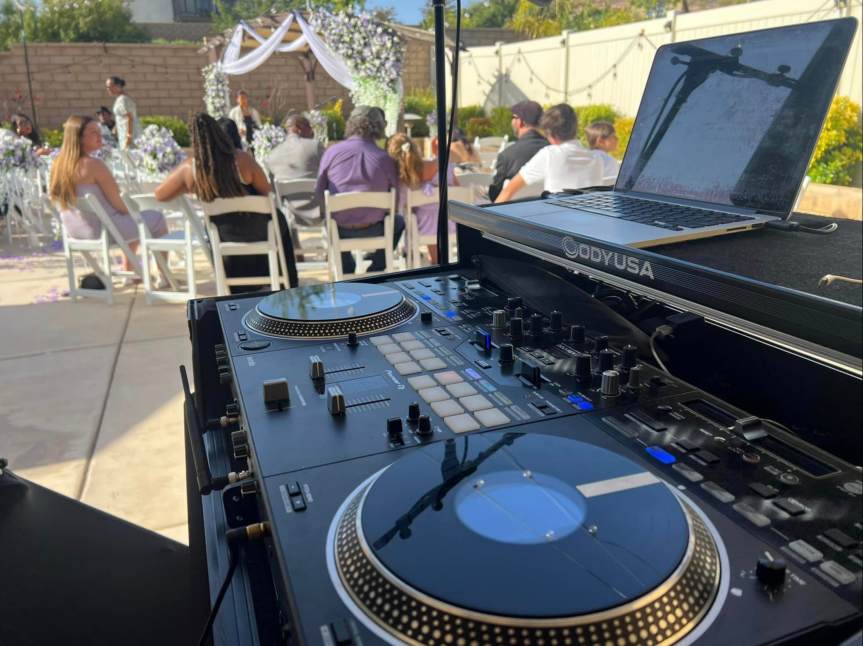 DJ setup at an outdoor wedding, with guests seated at tables in the background.