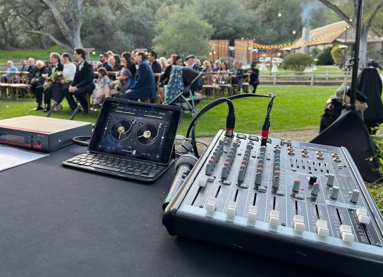 Audio mixing board and laptop set up for a wedding ceremony in a green field with guests seated in the background.