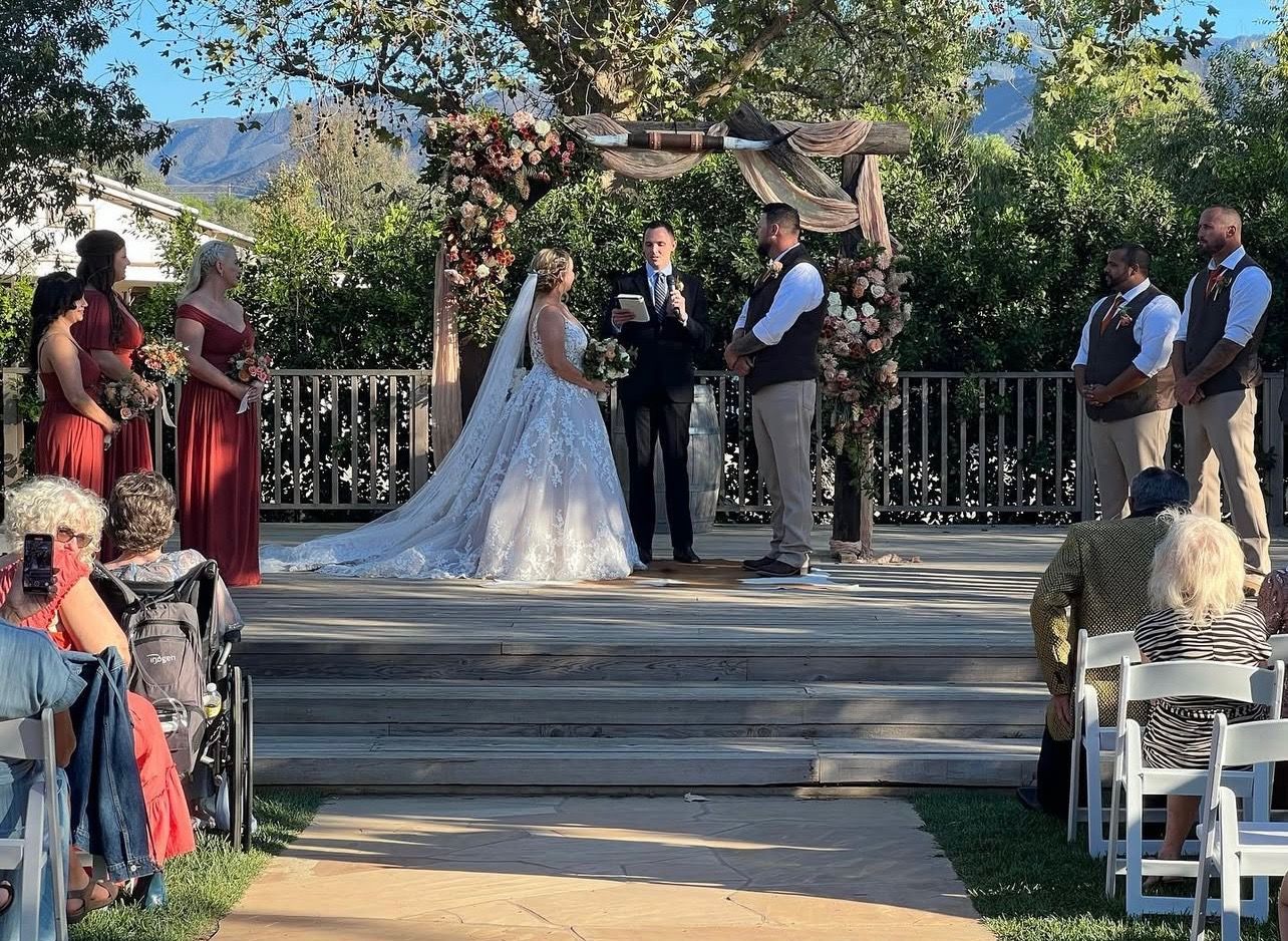 Wedding ceremony outdoors; bride, groom, officiant on stage, wedding party present, guests seated.
