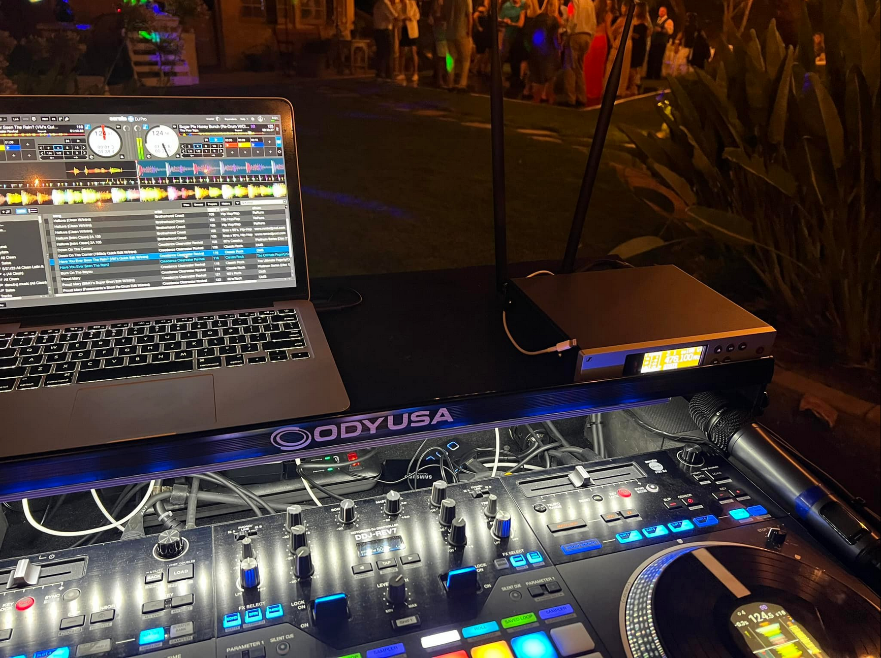 DJ setup at an outdoor event with a laptop, mixer, and people in the background.