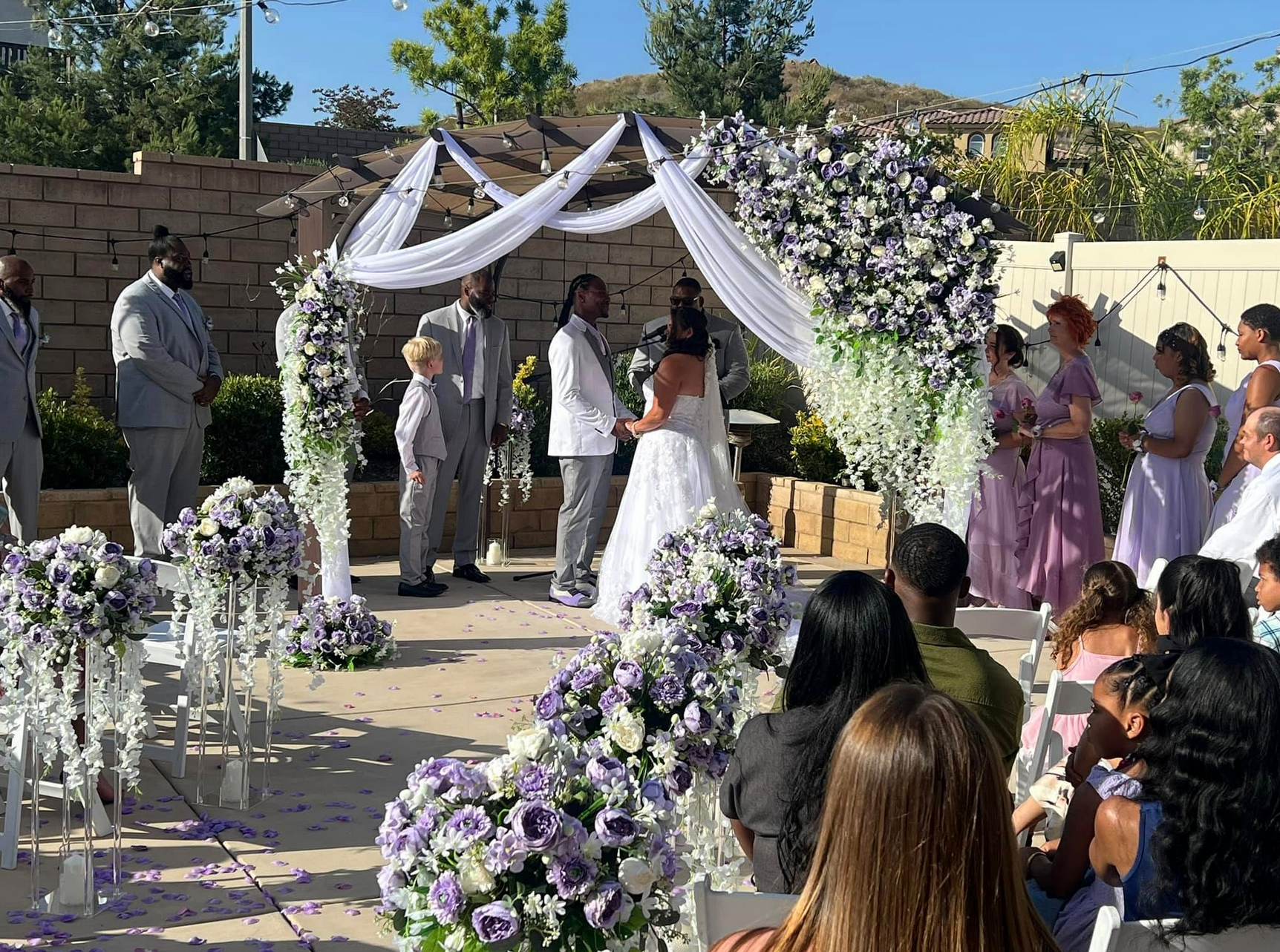 Wedding ceremony: couple under arch, surrounded by floral arrangements, guests seated outdoors.