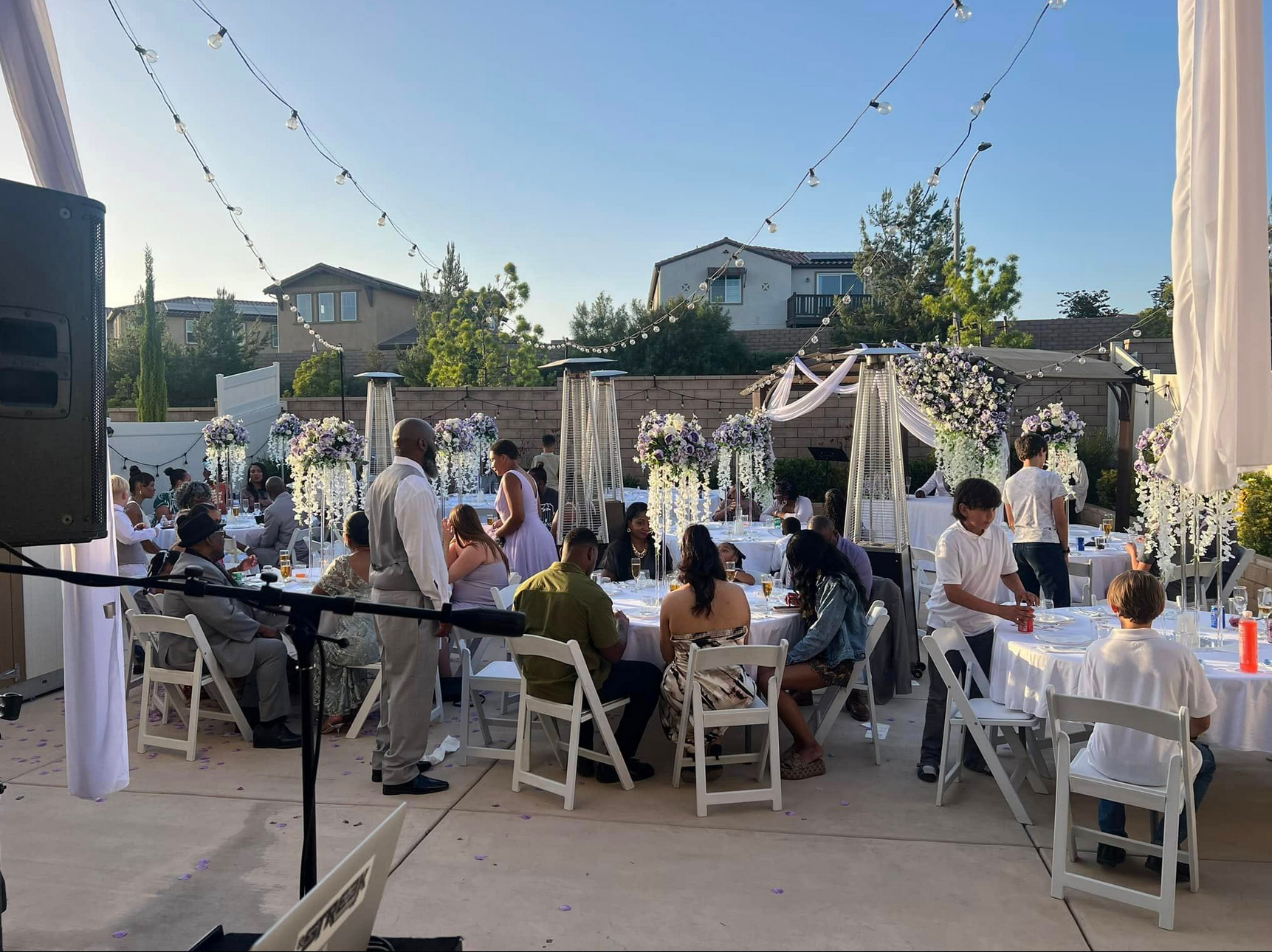 Outdoor party with guests seated at tables. Decorations, food, and music equipment are visible.
