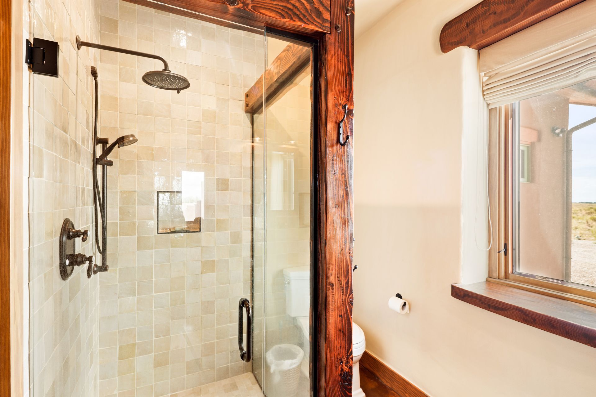 Glass shower stall with black fixtures beside a window in a bright bathroom with wood trim