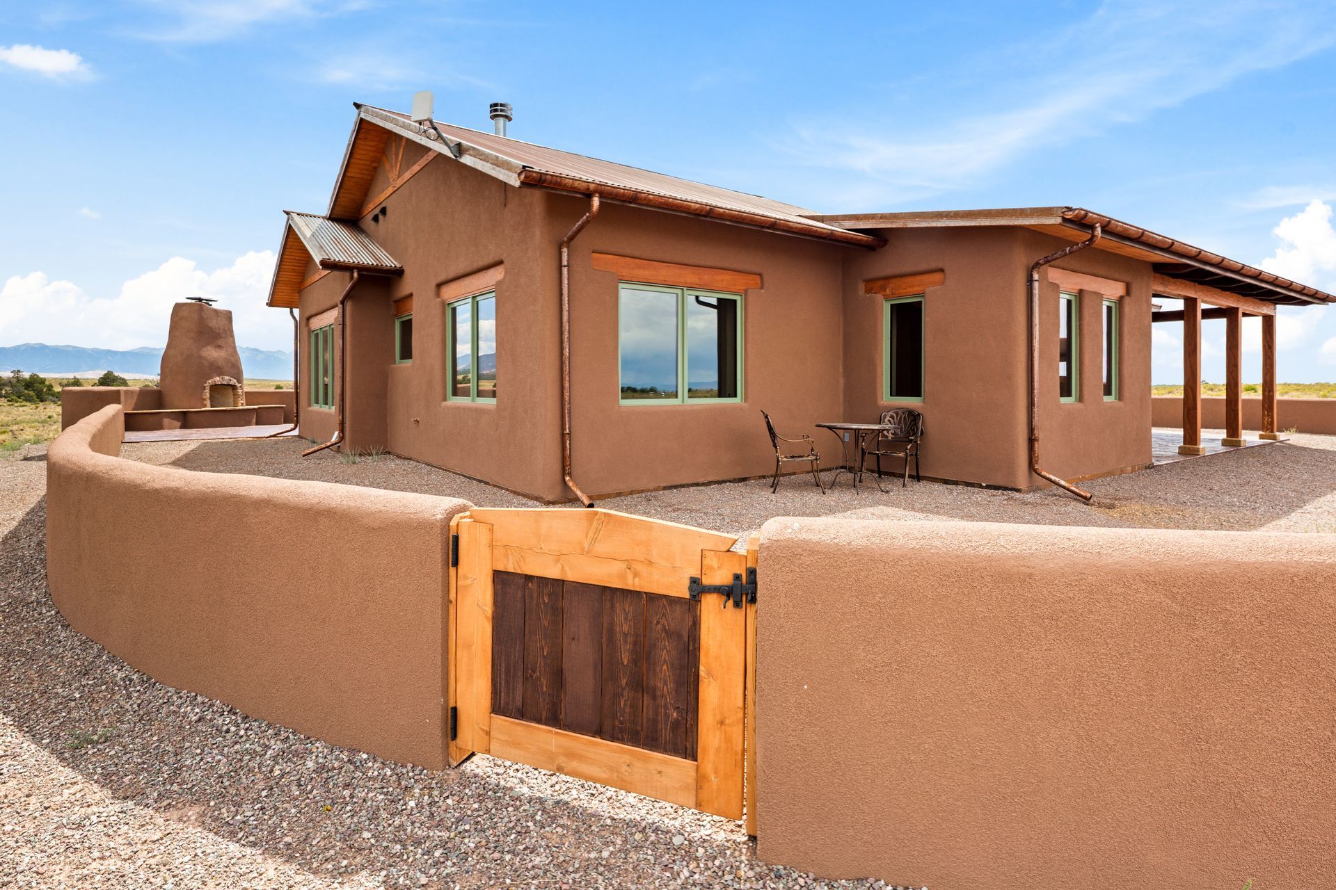 Adobe-style house with tan walls, wooden gate, and a low stucco courtyard wall under a blue sky