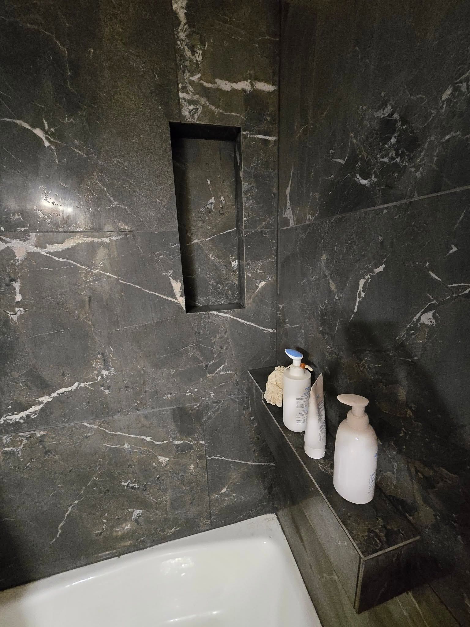 Black marble shower wall with recessed niche and bottles on a small ledge beside a tub.