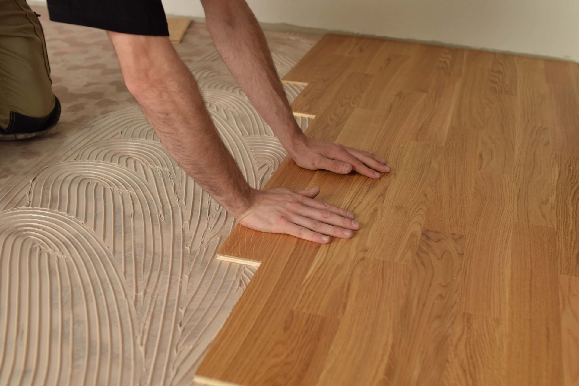 Hands and knees on a floor, installing a light wood laminate beside unfinished subflooring.