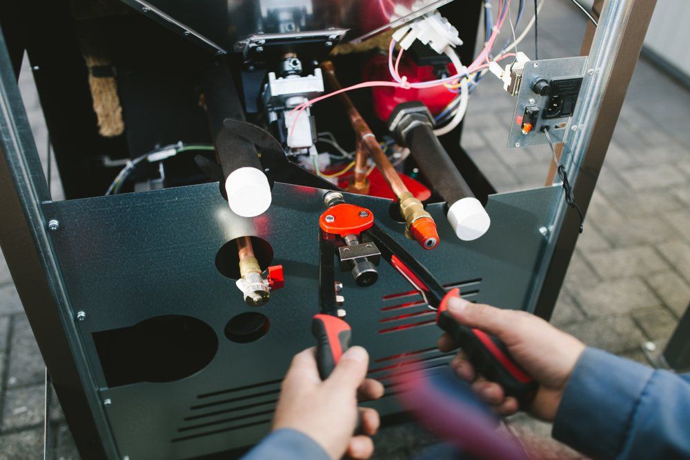 Hands using pliers to work on the inside of a furnace with exposed wiring and pipes.