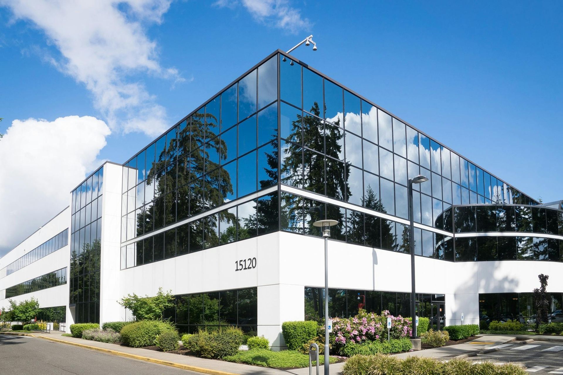 Modern glass-walled office building with white facade, blue sky reflection, and landscaping.